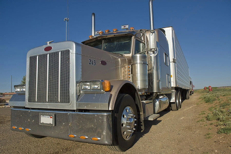 A classic beige semi-truck with a chrome front grill and long trailer parked along a dirt roadside.