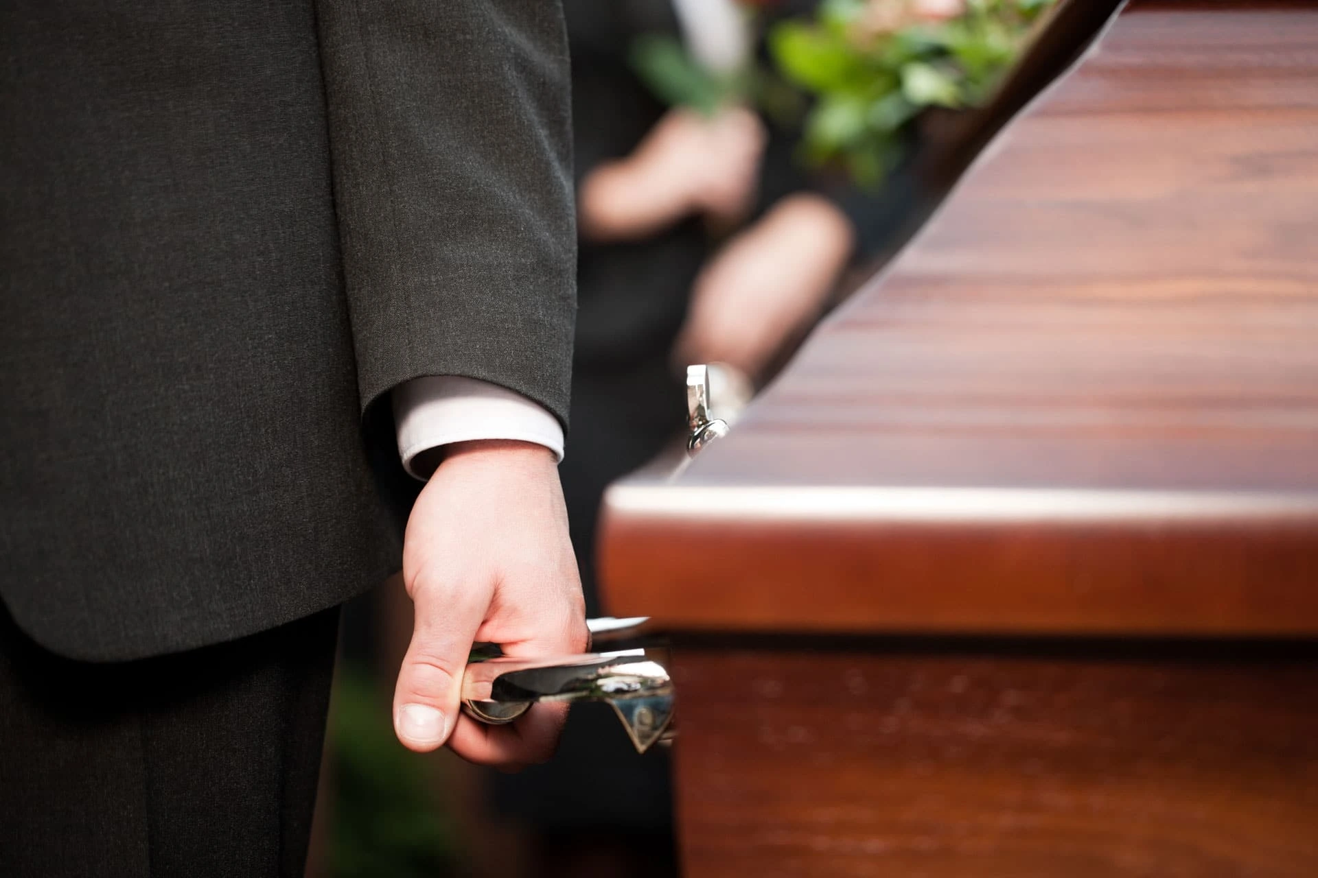 A close-up view of a pallbearer's hand carrying a wooden casket by its metal handle.