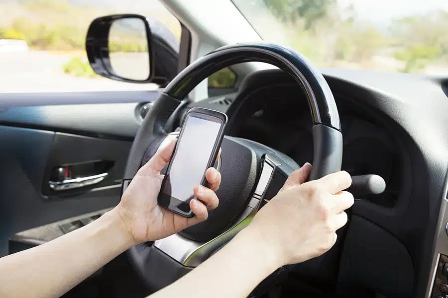 A driver holding a smartphone in one hand while gripping the steering wheel with the other.