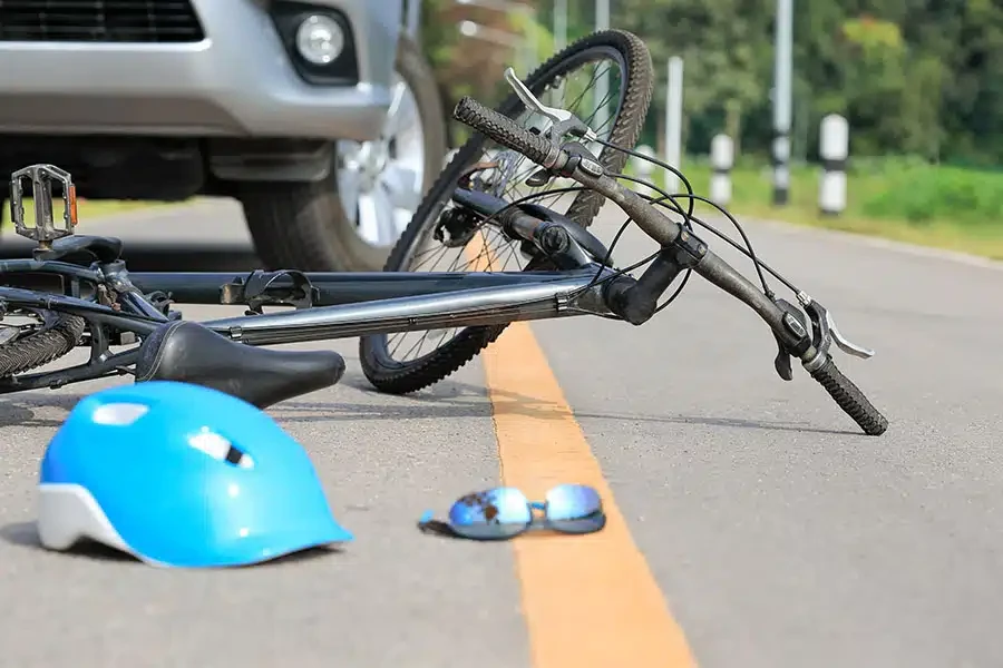 A bicycle lying on the road in front of a car with a blue helmet and sunglasses scattered nearby.