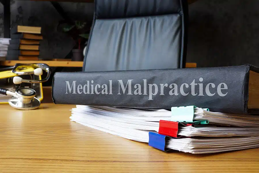 A thick black book with the title "Medical Malpractice" sits atop a stack of documents on a wooden desk, alongside a stethoscope and an office chair.