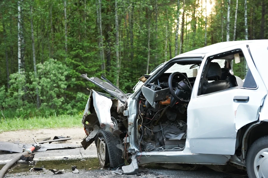 A heavily damaged car sitting on the side of a road near a forest after an accident.