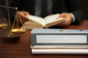 A gray binder sitting on books at a wooden desk with a scale of justice and a lawyer reading in the background.
