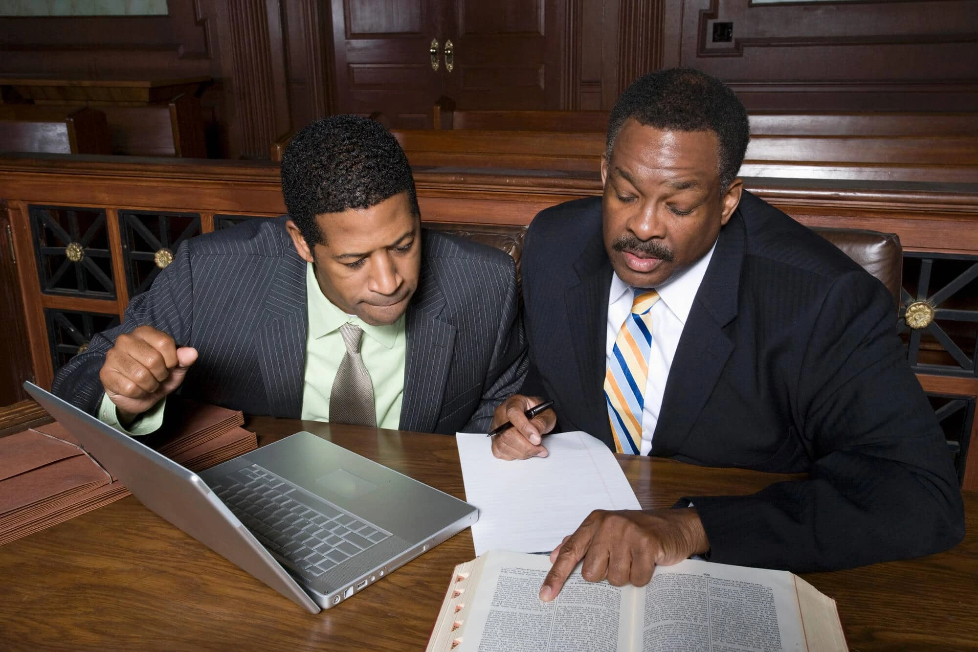 Two men in suits working together at a wooden table with a laptop, documents, and an open law book.