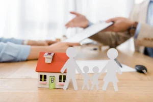 A toy house and paper cutout family figures sitting on a table while two people discuss documents in the background.