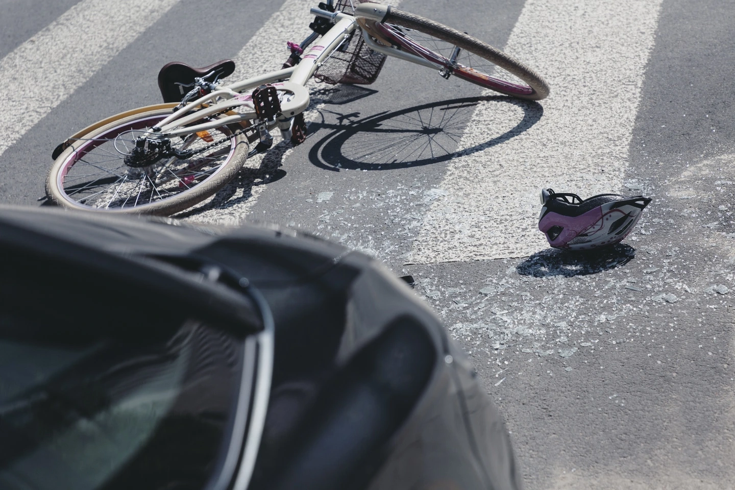 A bicycle lying on a crosswalk near a helmet and shattered glass in front of a car after an accident