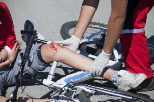 A paramedic wearing gloved hands attending to an injured cyclist's bleeding knee beside a fallen bicycle.
