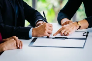 A close-up view of a person pointing to a document on a clipboard for another person in a suit to sign with a pen.