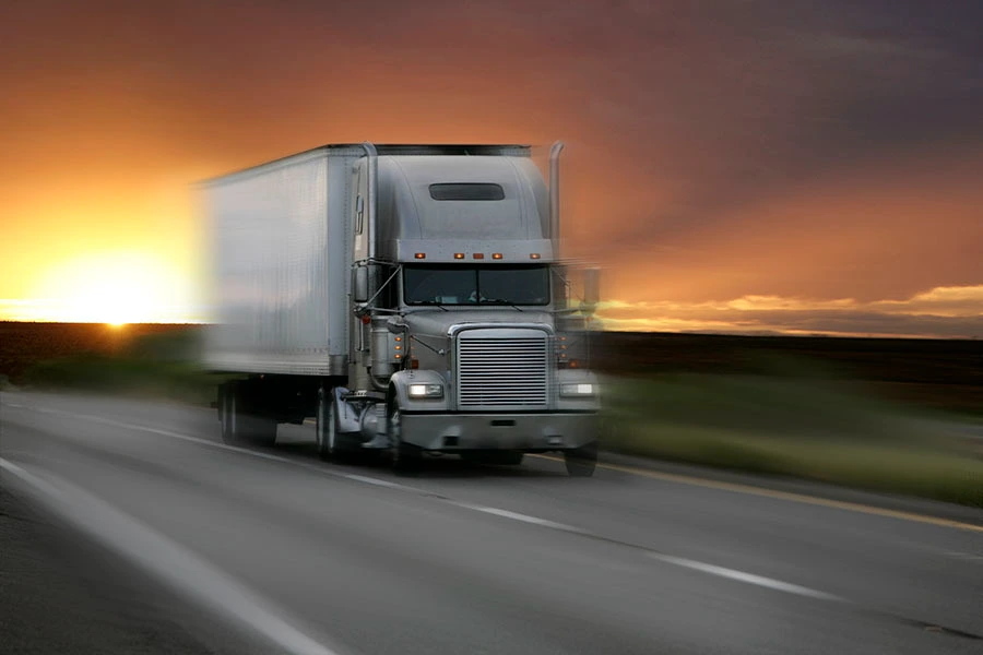 A silver semi-truck driving along a highway against a glowing sunset sky.