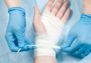 A pair of gloved hands applying a medical gauze bandage around a person's hand and wrist.