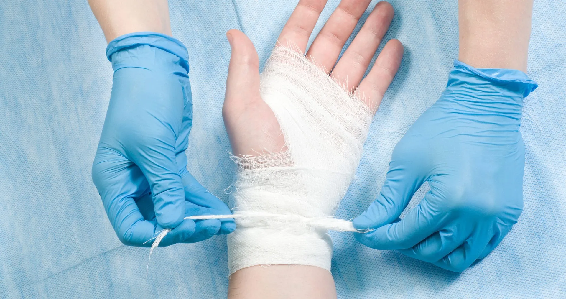 A pair of gloved hands applying a medical gauze bandage around a person's hand and wrist.
