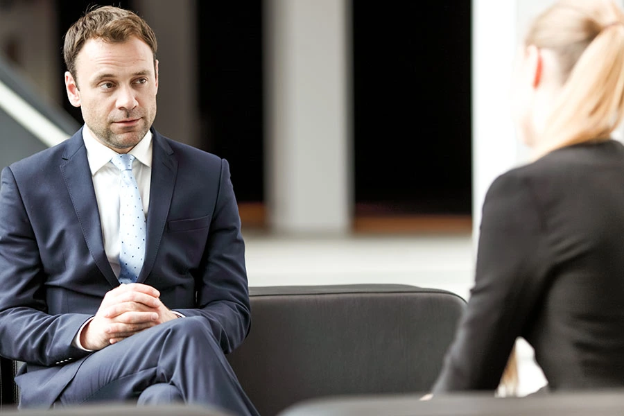 A man in a navy suit and light blue tie sits attentively across from a woman during a professional meeting or job interview in a modern office setting.