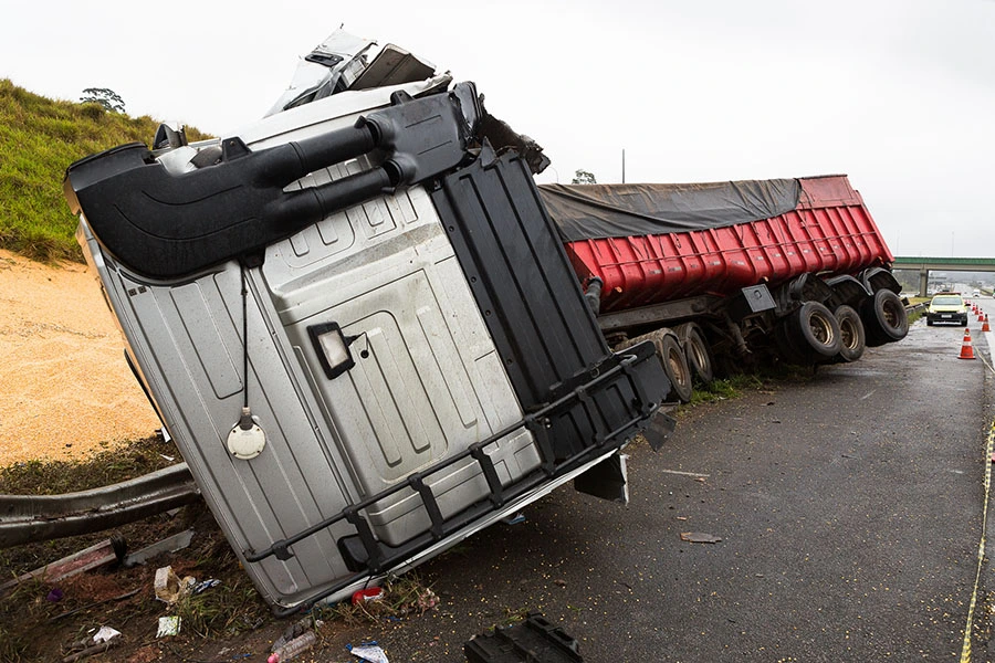 A crashed semi-truck with a silver cab and red trailer lying overturned on the side of a highway.
