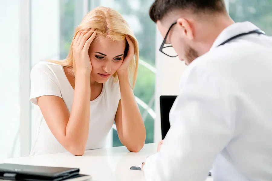 A stressed-looking woman holding her head in her hands while speaking with a doctor at a table.
