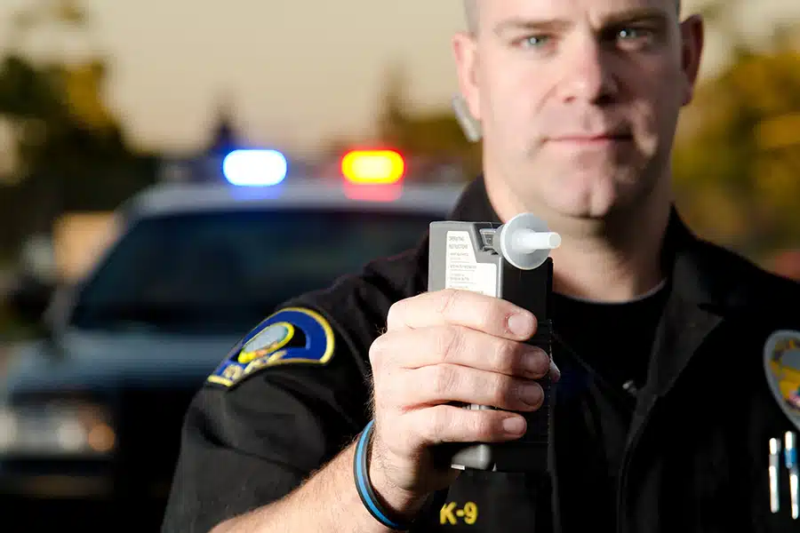 A police officer holding out a breathalyzer in front of a patrol car with flashing lights.