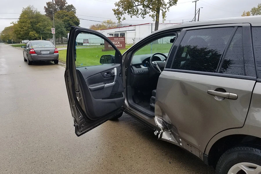 A grey SUV with damage to its lower side panel and driver's side door, parked with the front door open on a wet street.