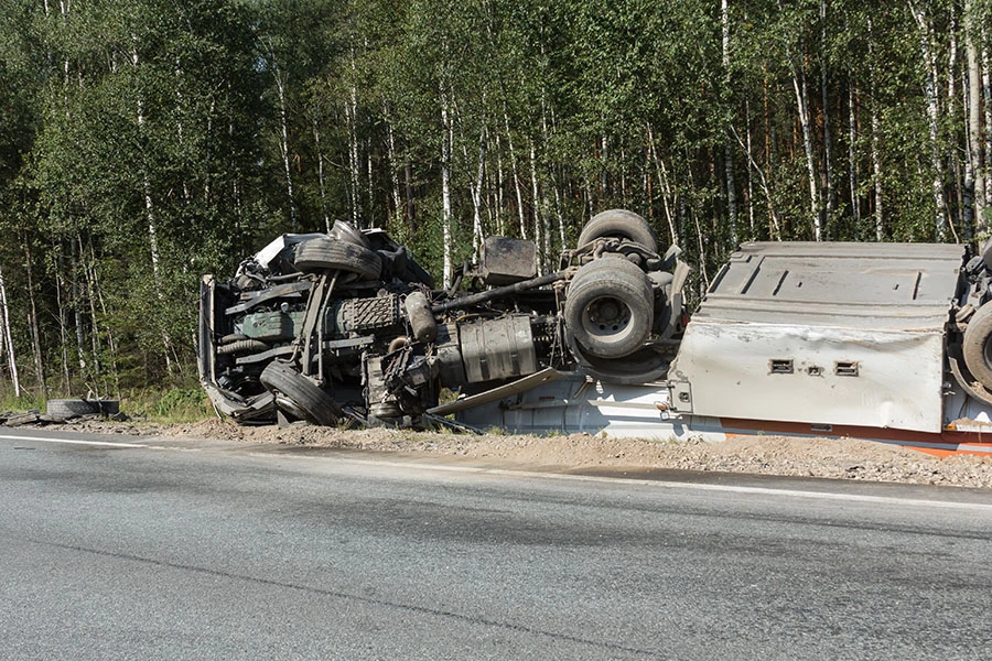 An overturned truck lying on its side along the edge of a road next to a forest.