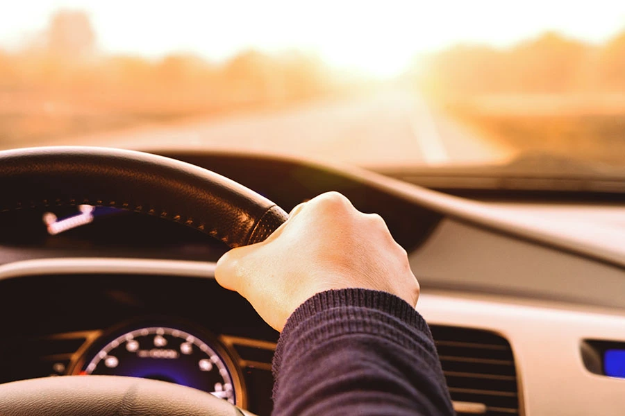 A driver's hand holding the steering wheel while driving toward the sunset.
