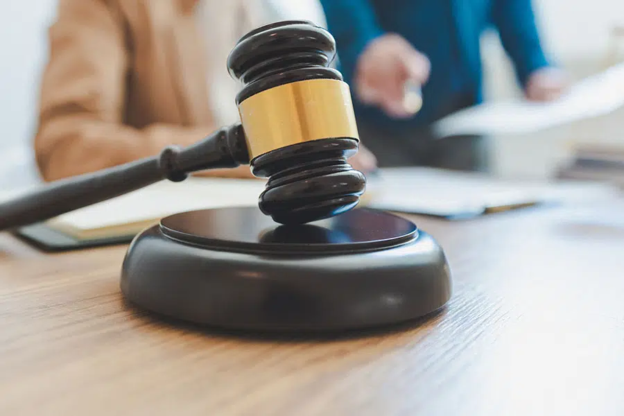 A judge's wooden gavel and sound block in close-up with people reviewing documents in the blurred background.