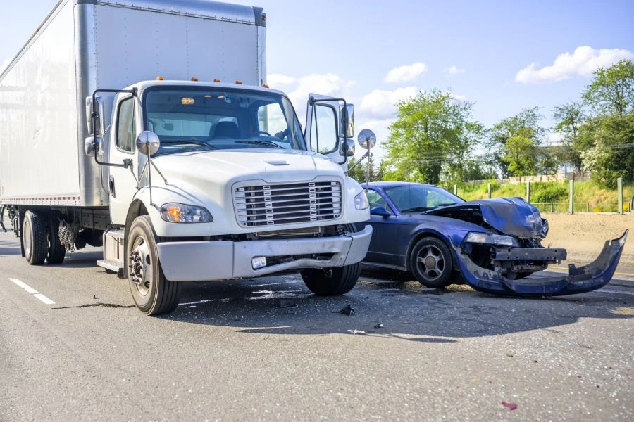 A blue sports car with severe front-end damage stopped next to a white box truck involved in a collision on a highway.