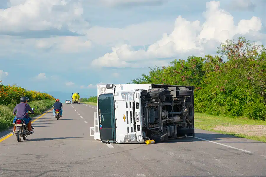 A small white box truck overturned on its side in the middle of a highway, with motorbikes passing by.