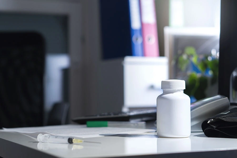 A white pill bottle and a medical syringe resting on a desk in a doctor's office or clinical setting.