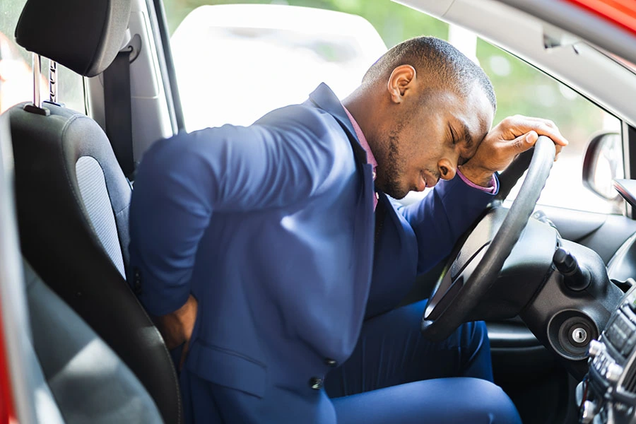 A man in a blue suit holding his lower back in pain while leaning over the steering wheel inside a car.