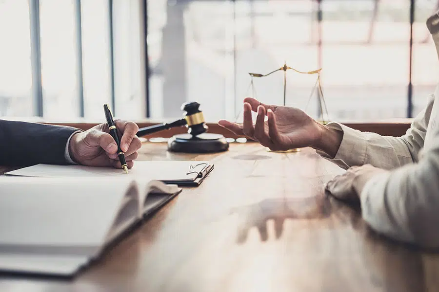 A client gesturing during a consultation with a lawyer taking notes at a desk with a gavel and scale of justice.