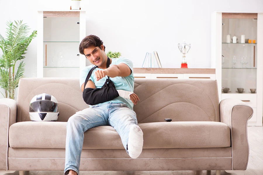 A injured man with an arm sling and casted leg sitting on a couch with a motorcycle helmet beside him.