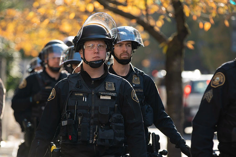 A line of Portland police officers in riot gear walking down a street with autumn trees in the background.