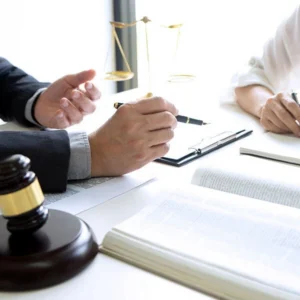 A close-up view of legal professionals discussing paperwork at a desk featuring a gavel, open law book, and scale of justice.