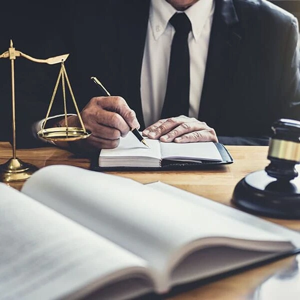 A lawyer writing in a notebook at a desk with a scale of justice, an open book, and a gavel.