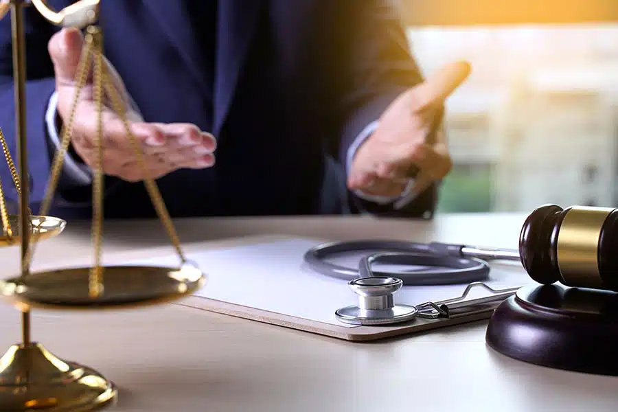 A person in a suit gesturing behind a desk with a stethoscope, a gavel, and a brass scale of justice.