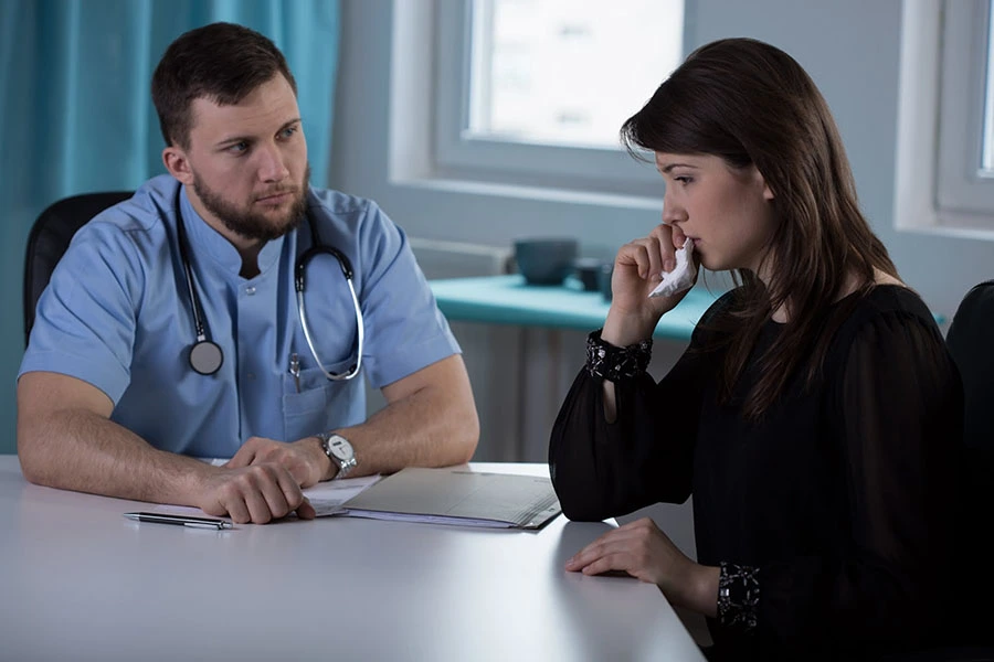 A male doctor in scrubs listening intently to a distressed female patient holding a tissue in a medical office.
