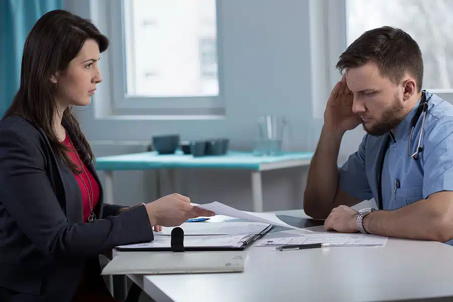 A stressed medical professional in scrubs consulting with a woman presenting documents across a desk.