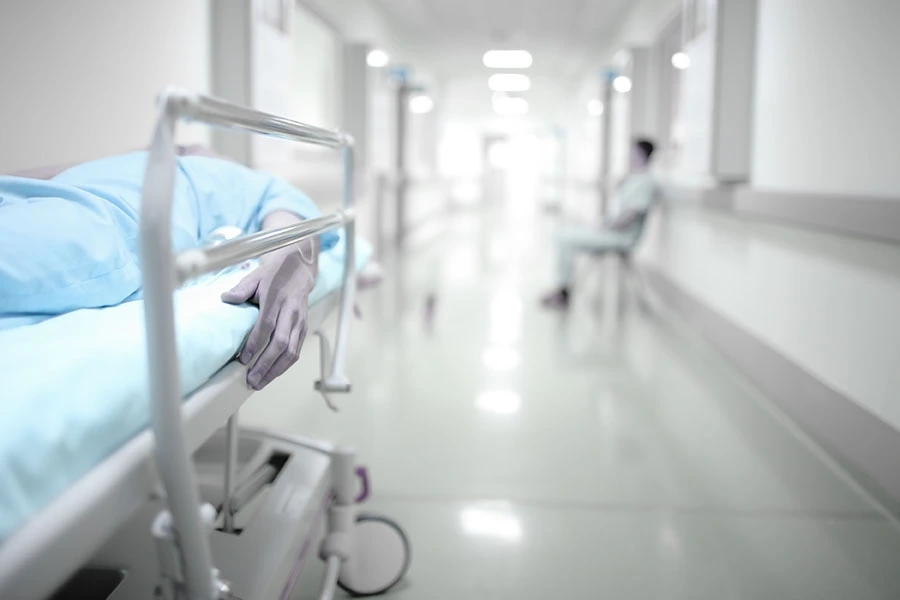 A patient lying on a hospital gurney in a bright, sterile corridor with a medical worker seated in the background.