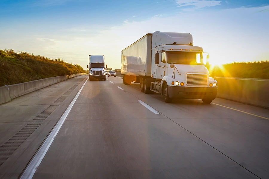 A white semi-truck driving along a highway during a bright, sunny sunset.