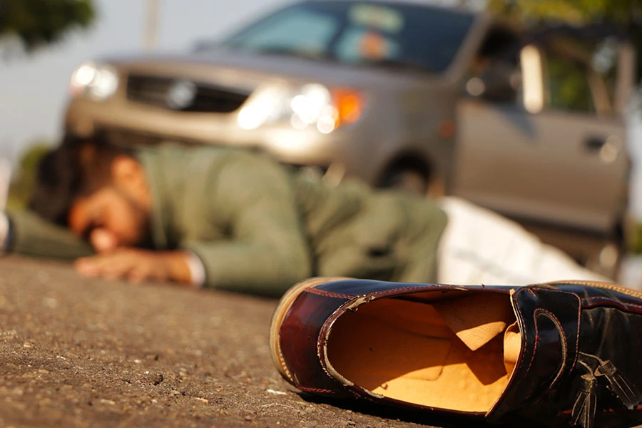 A shoe lying on the asphalt with an injured person face down and a car blurred in the background after an accident.