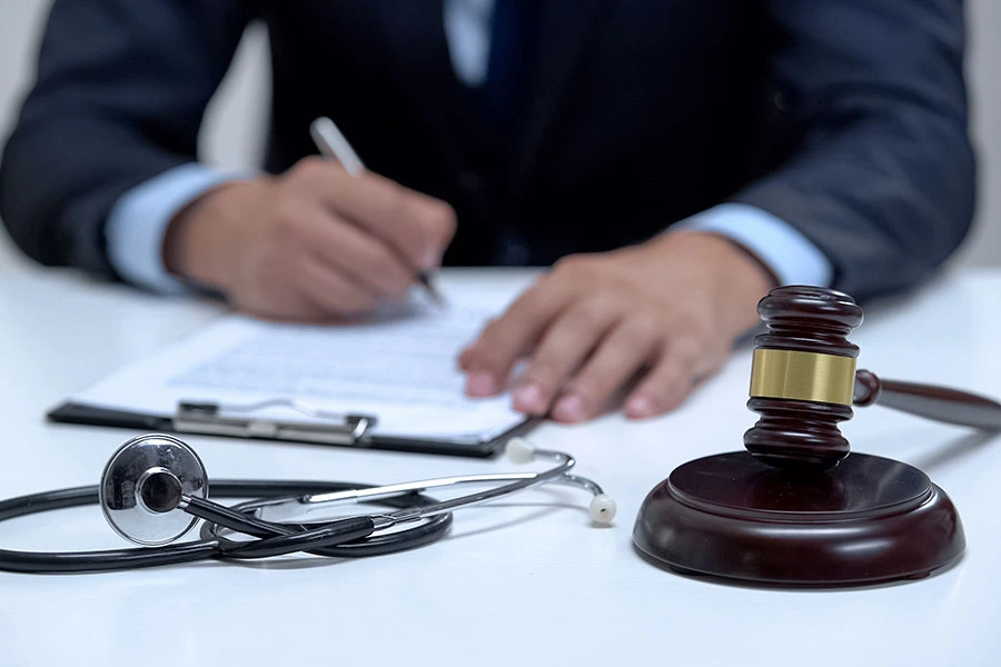 A judge's gavel and a stethoscope resting on a desk while a person in a suit writes on a clipboard in the background.