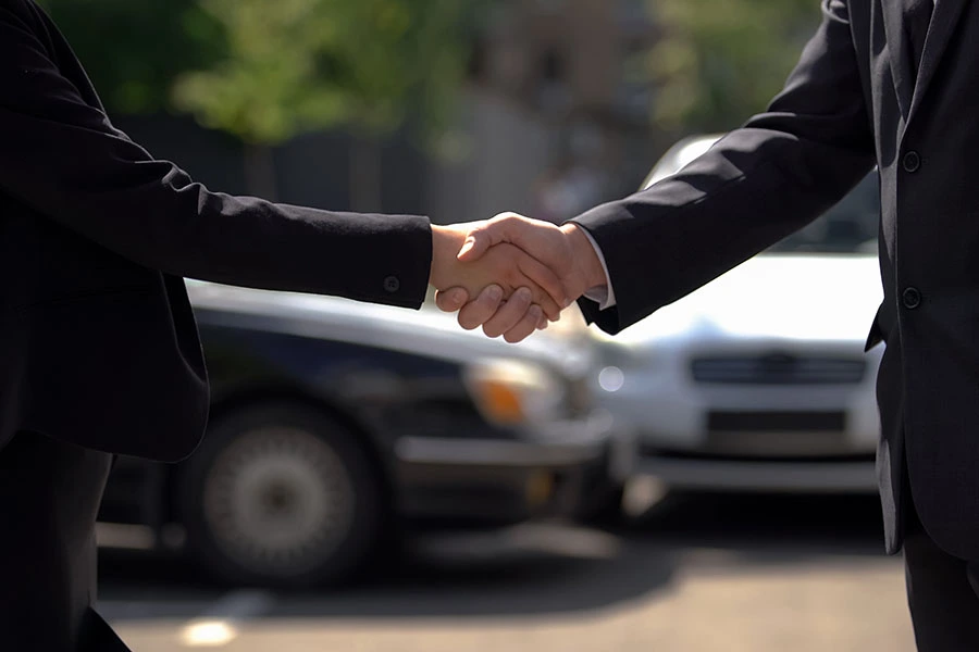 Two people in black suits shaking hands outdoors in front of two parked cars.