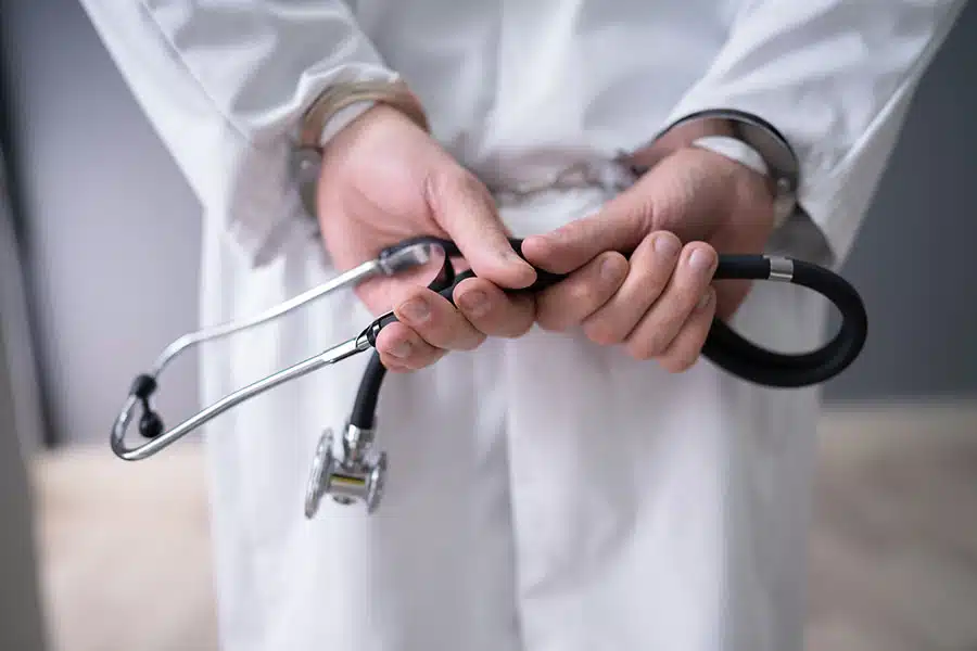 A doctor in a white lab coat holding a stethoscope with handcuffed wrists behind their back.