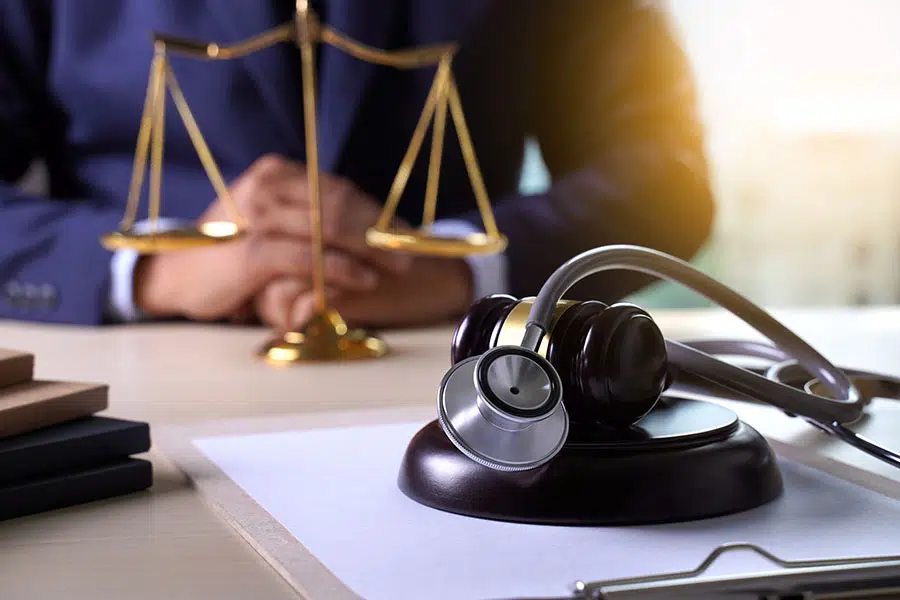 A stethoscope draped over a gavel on a clipboard, with a brass scale of justice and a person in a suit sitting in the background.