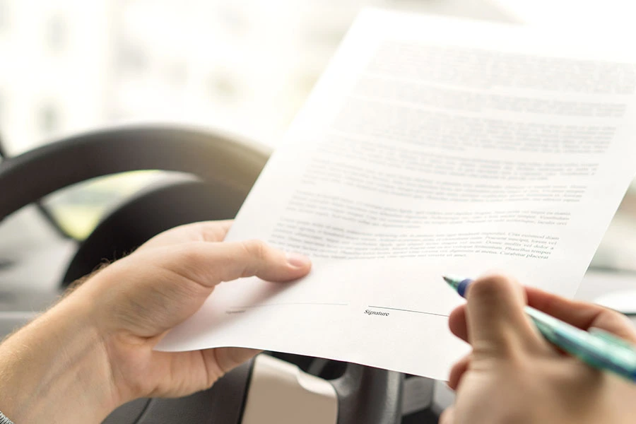 A person holding a pen, preparing to sign a document above the steering wheel inside a vehicle.