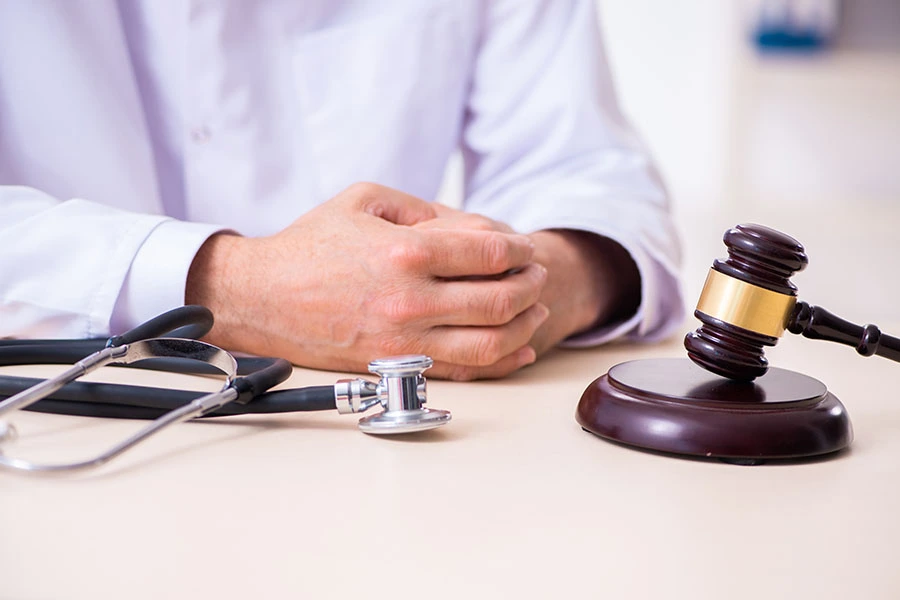 A doctor sitting at a desk with hands clasped, next to a stethoscope and a wooden gavel.