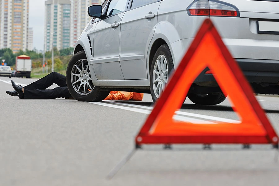 An warning triangle placed on the road with an injured person lying underneath a silver car following an accident.