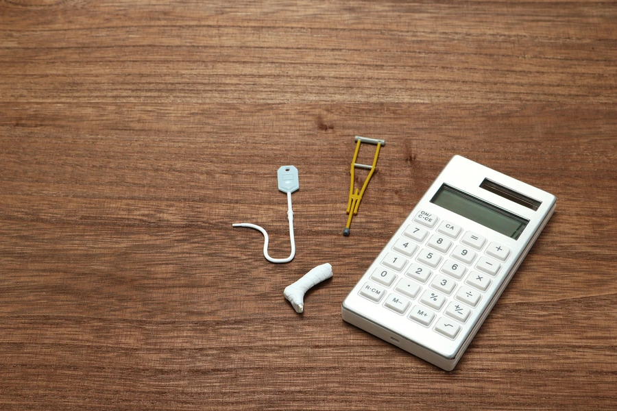 A white calculator placed on a wooden surface next to miniature medical objects including a crutch, a leg cast, and an IV bag.