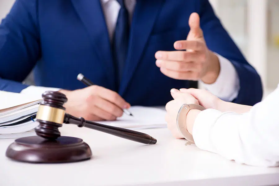 A lawyer in a suit speaking to a handcuffed client at a desk with a gavel in the foreground.
