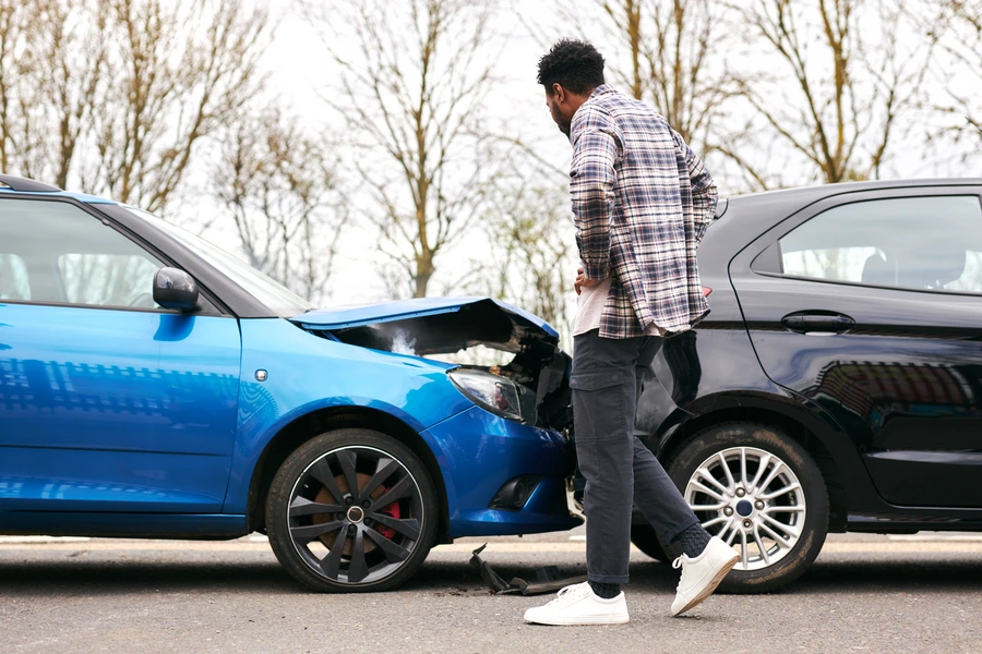 A man standing next to a blue car that has rear-ended a black car, looking at the smashed front hood.