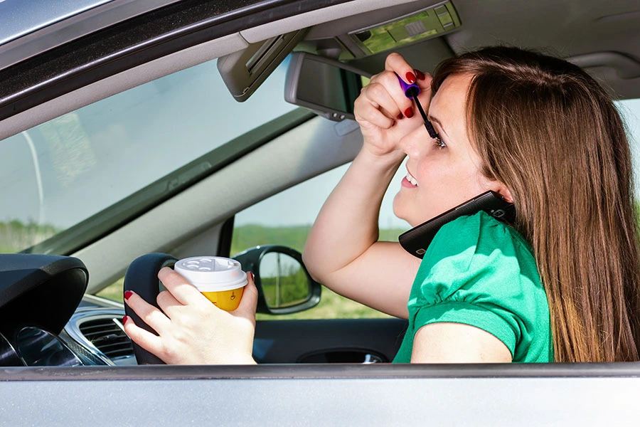 A woman applying mascara while driving, talking on the phone, and holding a cup of coffee.