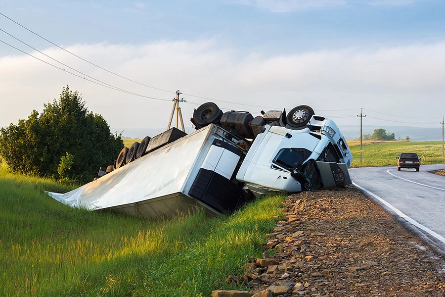 A white semi-truck and trailer overturned in a grassy ditch alongside a rural road.